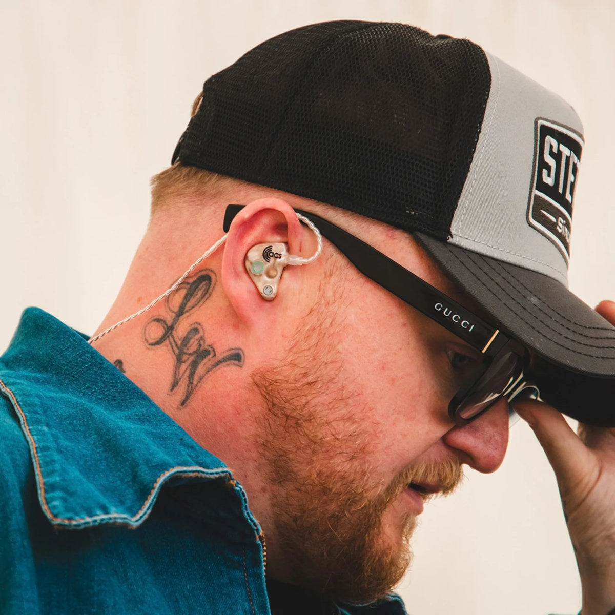 Man wearing a black and gray cap, sunglasses, and a blue shirt with a tattoo on his ear.