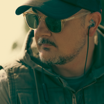 A close-up side profile of a man wearing a dark baseball cap, gold-framed sunglasses with dark lenses, and an in-ear earbud with a cord.