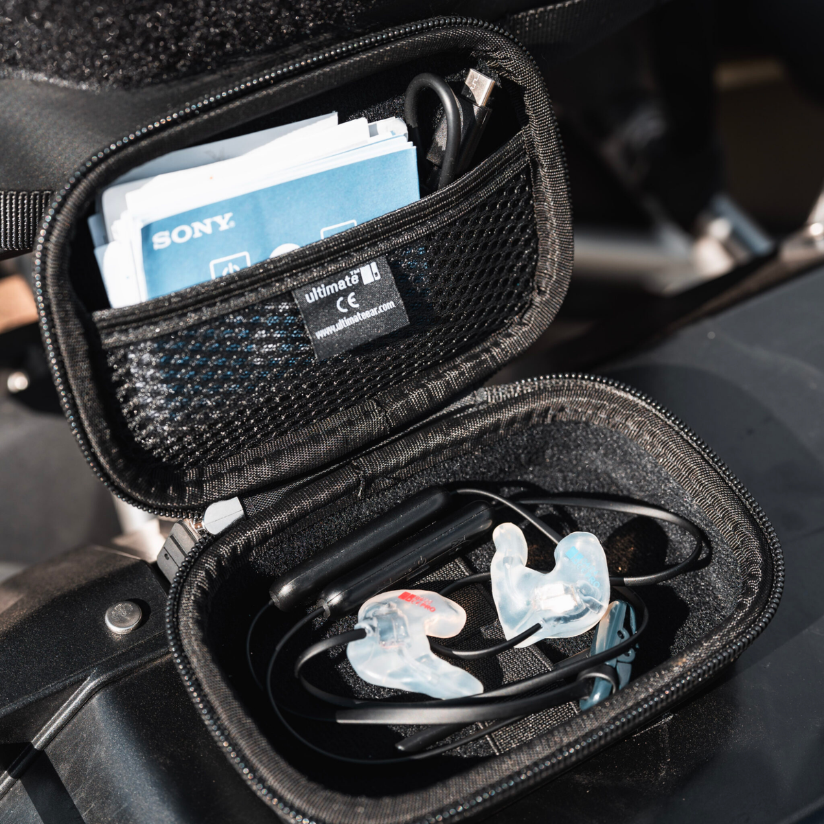 An open black zippered ear protection case containing clear silicone earplugs, a black strap, and a mesh pocket with a Sony card and cable, set against a dark motorcycle tank background.