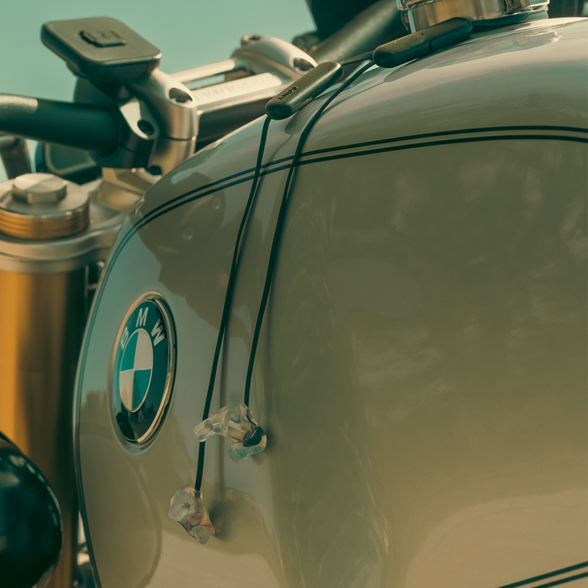 Close-up of a BMW motorcycle tank with the BMW roundel logo visible, showing part of the handlebar and fuel system against a softly blurred background.