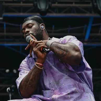 A man performing on stage, holding a microphone and wearing a light purple tie-dye shirt and matching head covering, with stage lights and equipment visible in the background.