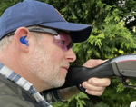 Man with ear protection and pink-tinted glasses holding a shotgun outdoors.