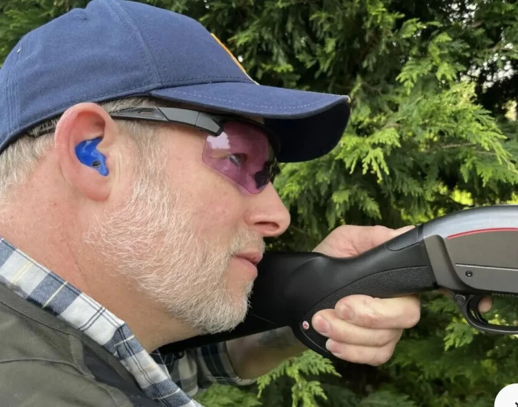 Man with ear protection and pink-tinted glasses holding a shotgun outdoors.