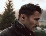 Man with a beard and earplugs outdoors with trees and mountains in the background