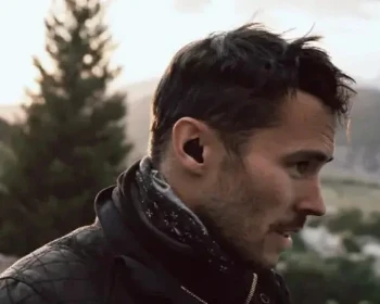 Man with a beard and earplugs outdoors with trees and mountains in the background