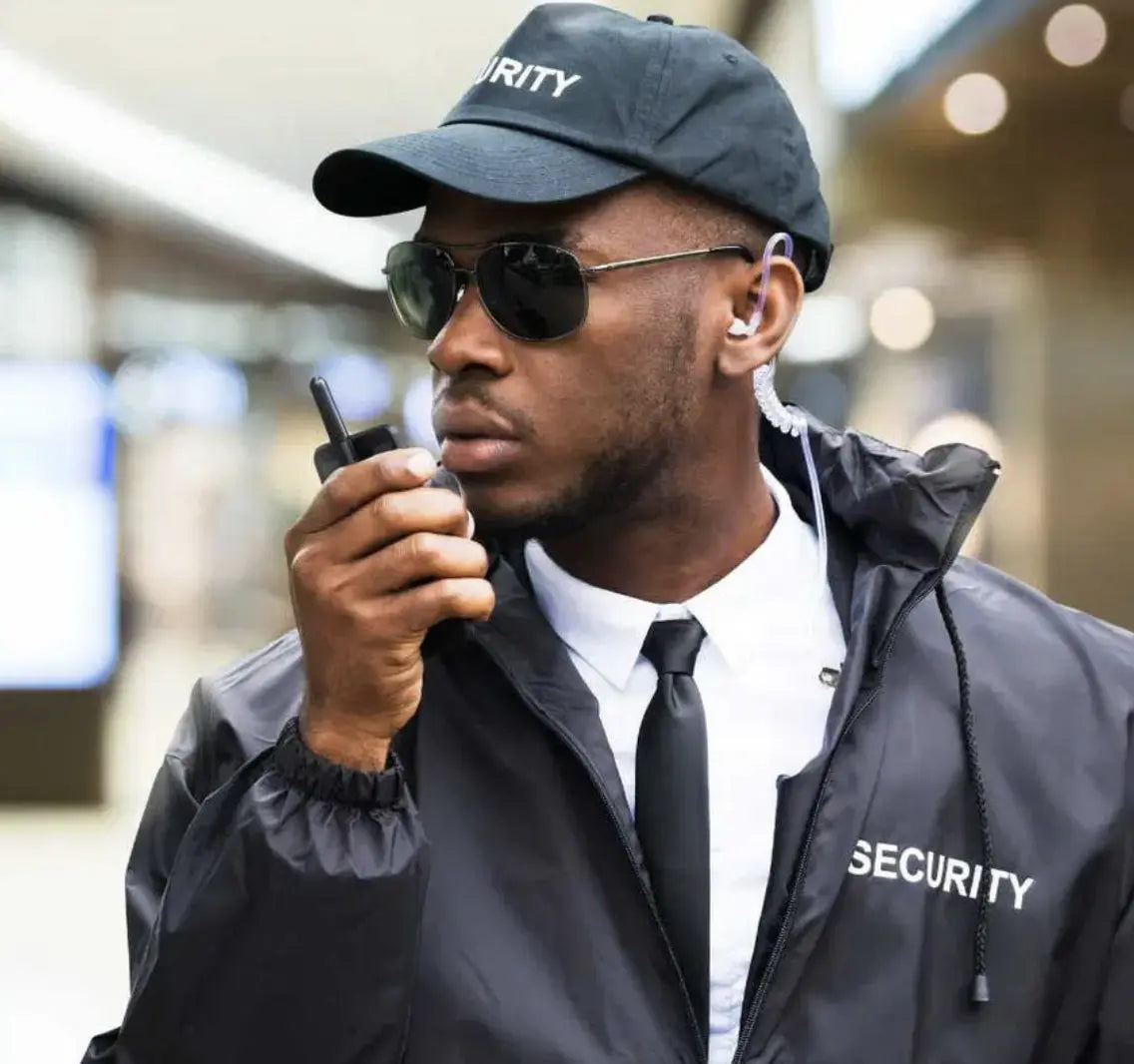 Security guard wearing a cap, sunglasses, and jacket with 'SECURITY' on it, holding a walkie-talkie.