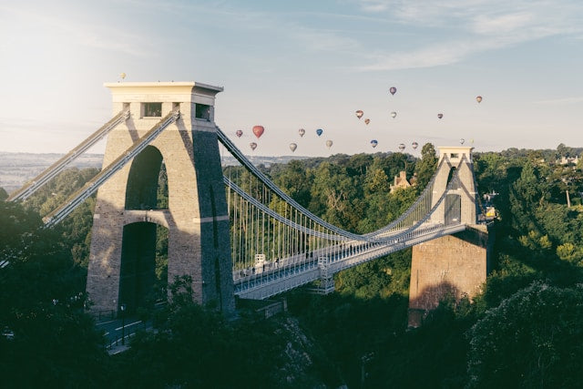 A scenic landscape photograph of the Clifton Suspension Bridge in Bristol, UK, with hot air balloons floating in the sky above a green forested valley.